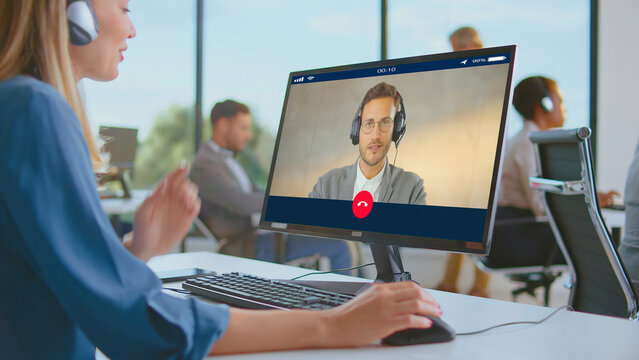 Professional woman wearing headset and working on desktop computer, engaging in an online video conference with male colleague in a contemporary open-plan office setting