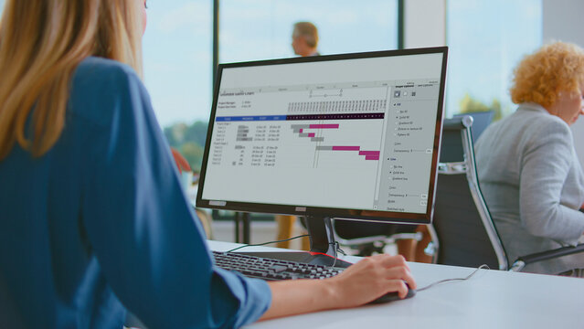 Businesswoman working on a computer screen displaying a project management gantt chart, planning tasks and tracking progress in a modern office environment with blurred colleagues in the background - Powered by Adobe