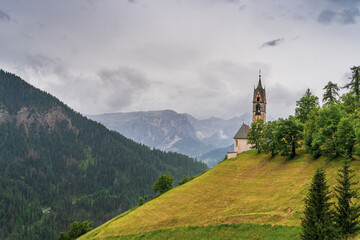 Fototapeta premium A dramatic summer storm sweeps over the Dolomite Alps, drenching the landscape in rain. Amidst the misty peaks, Chiesa di Santa Barbara stands graceful