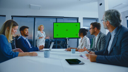 Diverse business professionals attending a corporate meeting in a modern conference room, watching a confident female speaker presenting on a green screen display board
