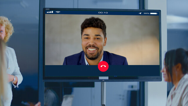 Diverse business team collaborating in a modern office during a video conference, watching a smiling manager on a large screen and engaging in remote teamwork and planning