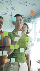 Professional businesswoman wearing glasses writing on glass board, marking strategic plan with sticky notes while colleagues brainstorming in background