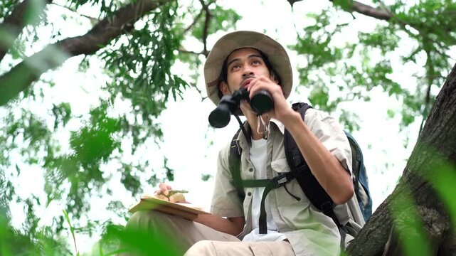 Asian male explorer observing birds with binoculars and taking notes.