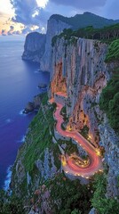 Cliffside road curves along the coast at dusk with lights illuminating the way