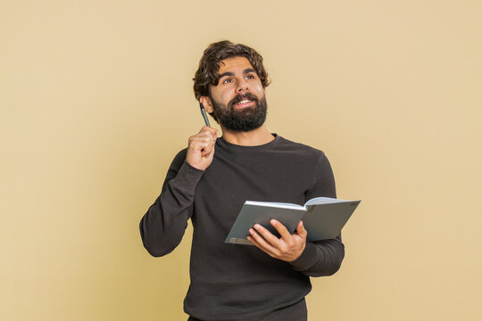 Thoughtful young Indian businessman making notes, writing down thoughts with pen into notepad notebook diary, to do list, good idea. Hispanic adult guy isolated on beige studio background indoors