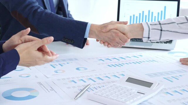 Business people are shaking hands and applauding, finalizing a deal while reviewing financial documents, graphs, calculator and charts on a desk in a modern office environment