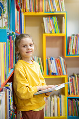 Portrait of a 7-8-year-old blonde girl in a yellow longsleeve in a library with books