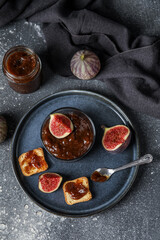 Plate with glass of sweet fig jam and tasty toasts on grey grunge background