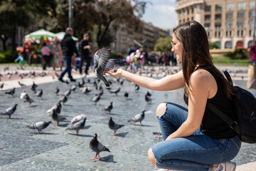 Cheerful young woman in Barcelona feeding pigeons in a city square, enjoying a beautiful sunny day and creating a lasting memory on her European vacation, travel adventure