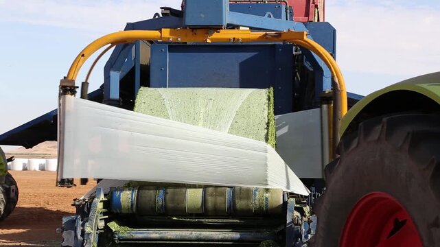 A bale wrapper is wrapping a bale of hay with plastic film in a field. The machine is blue and yellow. The bale is green. The field is brown. The sky is blue.