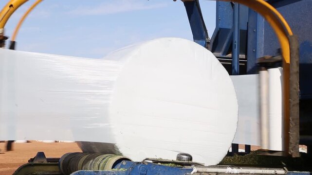 A bale wrapper is wrapping a bale of hay with plastic film in a field. The machine is blue and yellow. The bale is white. The field is brown. The sky is blue.