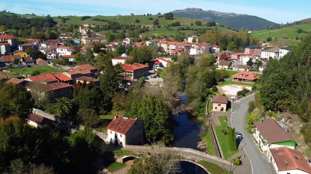 Aerial drone view of Lierganes, a historic village in Cantabria, Spain. The scene shows traditional stone houses, red-tiled rooftops, the medieval bridge over the Miera River, and lush green hills