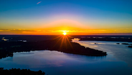 Aerial View of Sunset Over Dark Green Forested Land and Still Blue Waters Reflecting Orange Sky With Sunrays in Finland