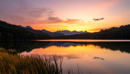 Fototapeta premium Golden Sunset Reflections on Tranquil Lake Surrounded by Lush Green Forest and Distant Mountain Range Casting Warm Glow over Calm Waters