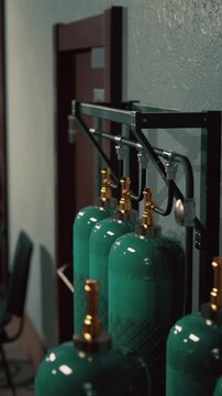 A row of green gas cylinders stands against a textured wall in a shadowy setting. Soft light highlights the bright valves, adding a touch of intrigue.