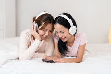 Two individuals lie on a bed, smiling and wearing over-ear headphones, sharing music. The cozy setting and joyful interaction highlight themes of friendship, bonding, and digital lifestyle at home.