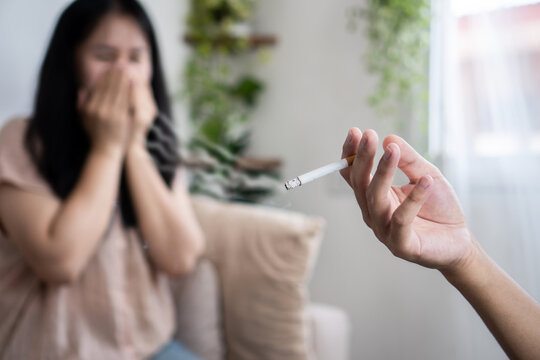 An Asian husband hand smokes next to his wife, who covers her nose to avoid the smell of cigarette smoke, demonstrating the harmful effects of secondhand smoke and bad habits at home.
