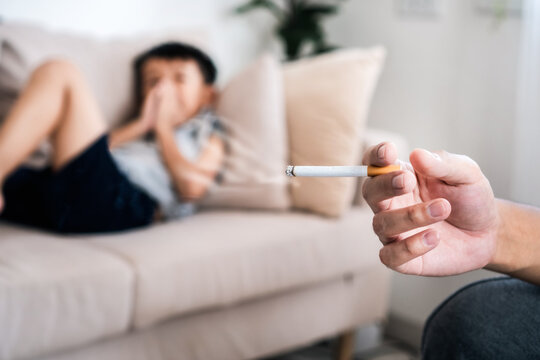 Asian father smoking next to his young son, The child covers his nose to avoid the cigarette smell, showing the dangerous effects of secondhand smoke and bad habits at home. 
