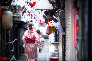 Elegant woman in a traditional kimono strolls through a charming, decorated Japanese alleyway with lanterns and white sakura blossoms. Cultural Japan travel, traditional clothing, Asian street.