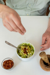 Hand adding salt and chilli to guacamole (avocado) in a white bowl. Raw Food on a white table. Chilli flakes and mortar (Raw Spice) present. Healthy recipe.