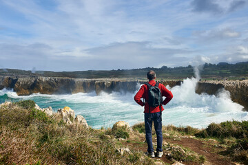 A man in a red jacket watches visitors walking above the cliffs of Pria with waves crashing against the rocks