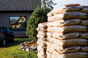 A stack of 15 kg wood pellet bags in the foreground, a house with a dark tiled roof and large window, a black car, and a small evergreen tree visible.