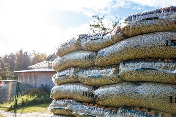 Bags of wood pellets are stacked outdoors, wrapped in clear plastic. A wooden structure with a slanted roof and trees are visible under a sunny sky.
