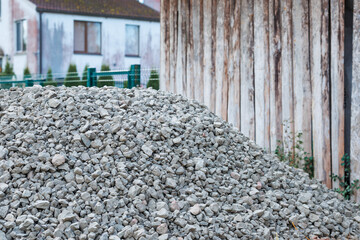 A pile of gray gravel sits near a weathered wooden structure, with residential buildings and tiled roofs visible behind a green metal fence.