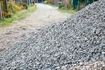 A large gray gravel pile sits on a dirt pathway bordered by grass, fencing, and plants. Residential structures and trash bins are visible along the path.