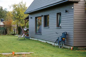 A gray wooden house with a sloped roof, three windows, a bicycle with child seat, hammock, folding chair, grass, trees, and a fence in the distance.