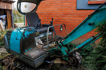 A teal and black excavator with rust and peeling paint sits near a red brick wall. A wooden shed with materials is visible, surrounded by vegetation.