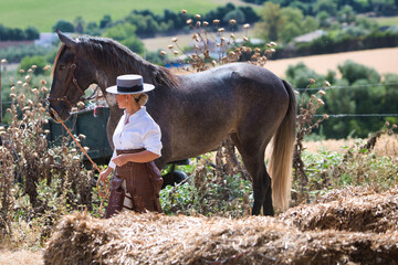 A young, beautiful woman riding a horse, dressed in riding clothes and a typical Spanish hat, is walking alongside her grey horse in the countryside.