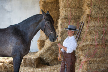 A young, beautiful woman dressed in riding clothes and a traditional Spanish hat is standing next to her grey horse in the barn, preparing to ride. The woman is in love with her horse.