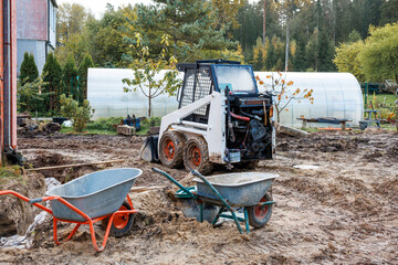 A skid steer loader on muddy ground, two wheelbarrows in the foreground, a white greenhouse, young trees, and a red walled building near a forested area.