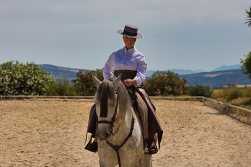 A young, beautiful Spanish woman dressed in riding clothes and wearing a traditional Spanish hat, riding a horse in the countryside on a sunny day.