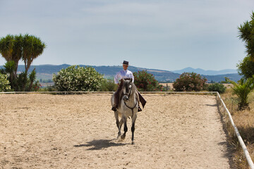 A young, beautiful Spanish woman dressed in riding clothes and wearing a traditional Spanish hat, riding a horse in the countryside on a sunny day.