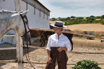A young, beautiful Spanish woman dressed in riding clothes and wearing a traditional Spanish hat stands next to a horse in the countryside on a sunny day. More horses graze in the background.