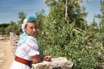 A beautiful young woman wearing a traditional white dress from Andalusia for flamenco dancing and a flower in her hair is holding onto a wooden fence. Olive trees in the background.