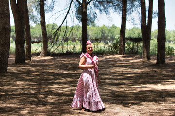 A beautiful young Spanish woman dressed in a traditional pink and white flamenco dress with a flower in her hair walks among the pine trees of Andalusia during the pilgrimage.