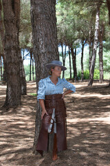A young, pretty woman dressed in riding breeches and riding clothes and a typical Spanish hat rests, leaning against a pine tree in the shade on a pilgrimage day in Spain.