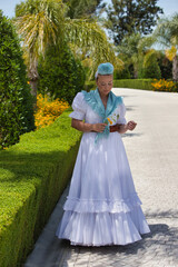 A beautiful young Spanish woman dressed in a traditional white Andalusian flamenco dress with a flower in her hair. The woman picks up a yellow flower with her hand and poses for the photo.