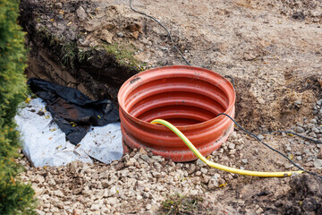 An orange corrugated pipe partially embedded in gravel and soil, with a yellow hose and black cable extending into it near a partially dug trench.