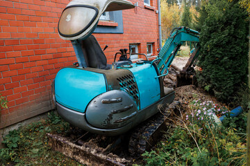 A weathered blue excavator sits by a red brick building with visible windows, surrounded by overgrown plants, soil, a bush, and flowers.