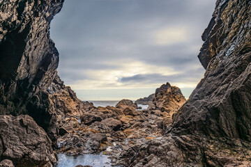 Rugged Irish shoreline seen through towering cliffs and boulders under a moody overcast sky.