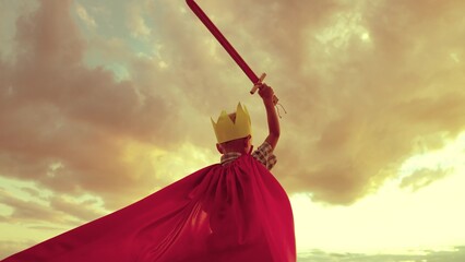 boy plays superhero against backdrop sunset sky. small child kid red cape with sword his hand....