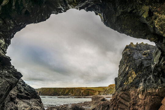 Dramatic sae arch rock-framed Irish coastline with turbulent sea, small beach, and distant cliffs under clouds.