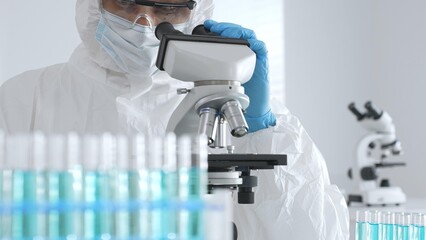 Male doctor in protective suit and gloves using microscope while analyzing samples while conducting research in sterile laboratory. Close-up of test tubes with blue liquid