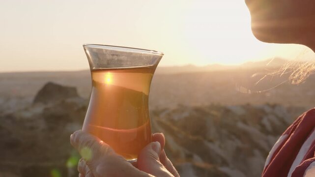 Turkish tea in traditional glass in sunlit sunset in hands of woman against backdrop of mountain landscape close-up