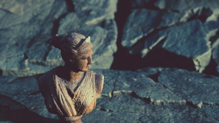 A detailed bronze bust stands proudly on rough stone surfaces. Natural light highlights the texture and detail, creating a serene yet striking scene by the outdoors.