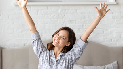 A happy young woman in pajamas is stretching her arms above her head in bed, waking up feeling refreshed. - Powered by Adobe
